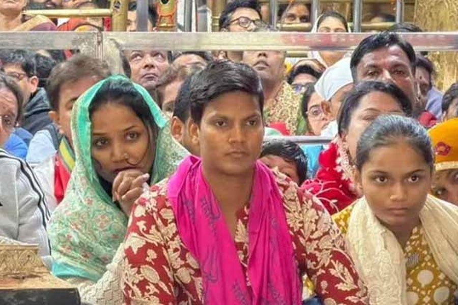 Pink name necklace around his neck. 'Mahadeva' written on it. Short hair. Temple prayers, shaking hands in rhythm with bells and chimes. Tattoo all over his forehead.